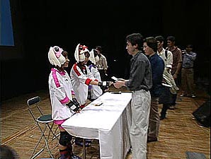 The three voice actors in cosplay outfits greet fans behind a table.